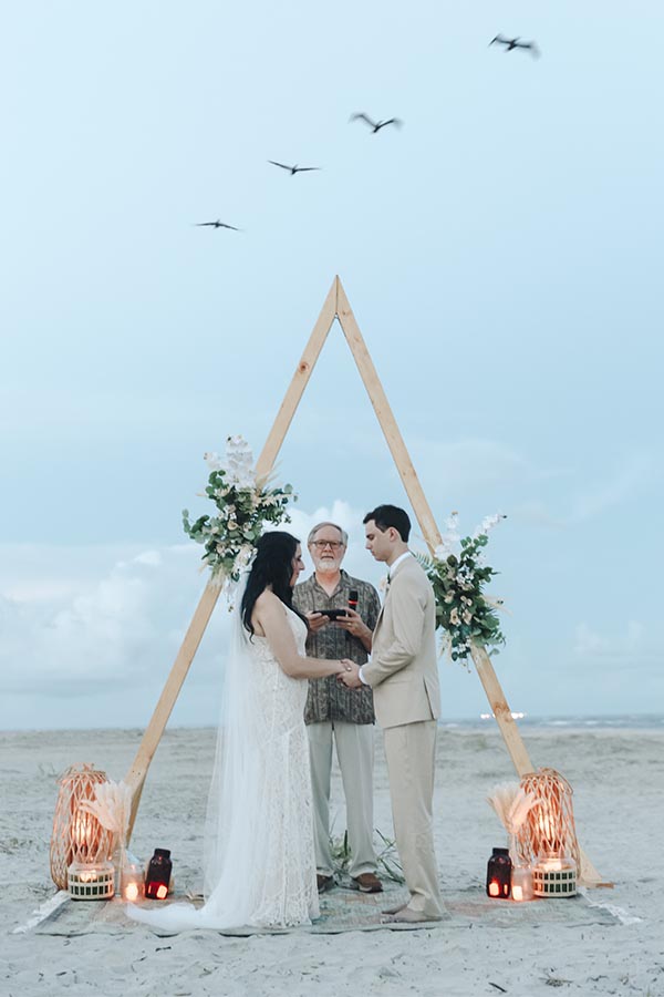 Bride and groom exchanging vows on the beach under wooden triangle arch during intimate elopement ceremony