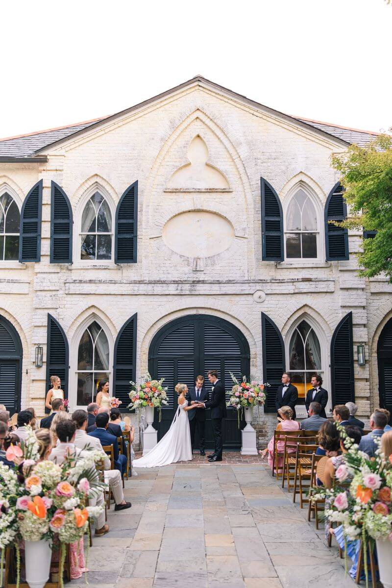 Bride and groom exchanging vows during their outdoor wedding ceremony at Alhambra Hall in Mount Pleasant, SC, surrounded by guests and vibrant orange and pink floral arrangements