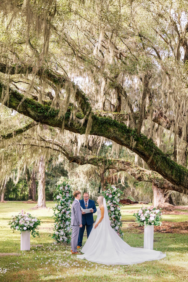 Bride and groom exchanging vows with an officiant under a giant live oak with Spanish moss and white floral pillars, Charleston elopement