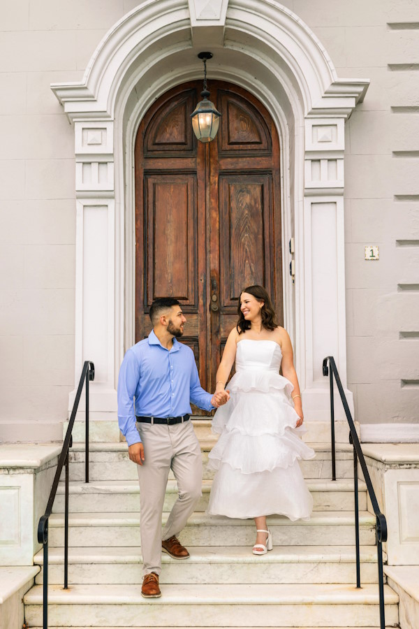 Engaged couple holding hands and walking down the front steps of a historic Charleston home with an arched wooden door