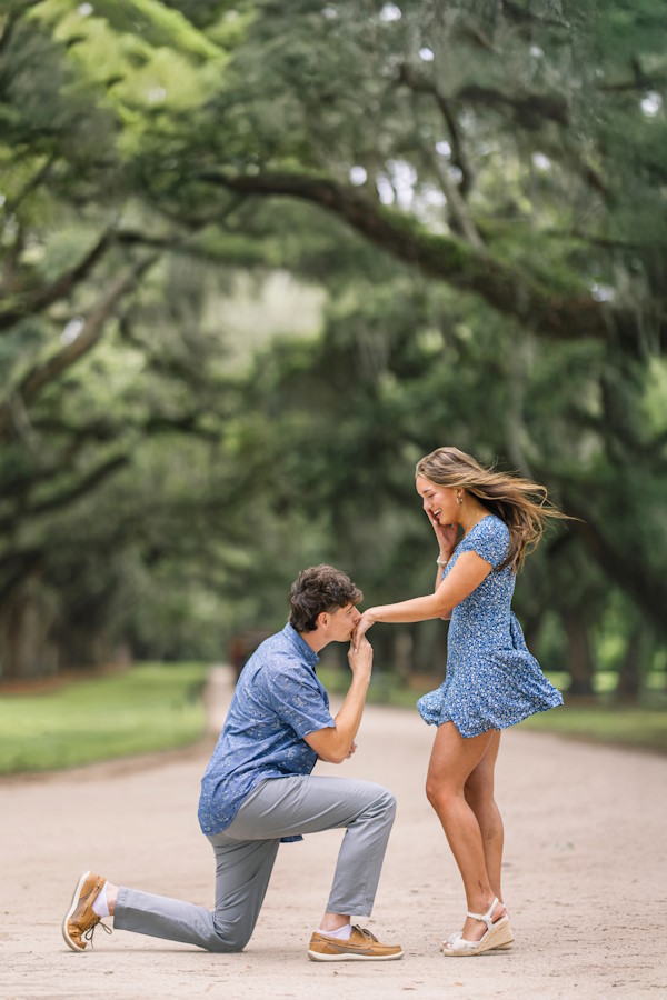Man on one knee kissing his girlfriend’s hand during a surprise proposal under moss-draped live oaks near Charleston, SC