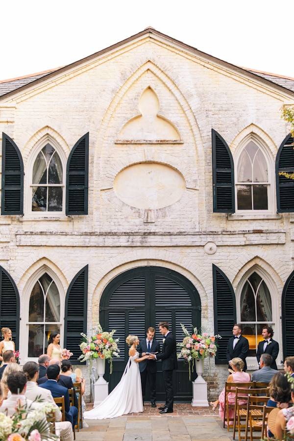 Outdoor wedding ceremony in the courtyard of the William Aiken House in Charleston, SC, with pink and coral floral arrangements along the aisle